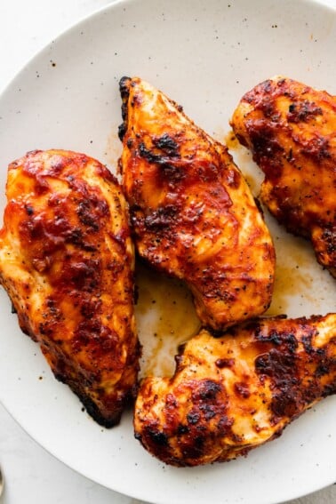 Four grilled BBQ chicken breasts on a white serving plate.
