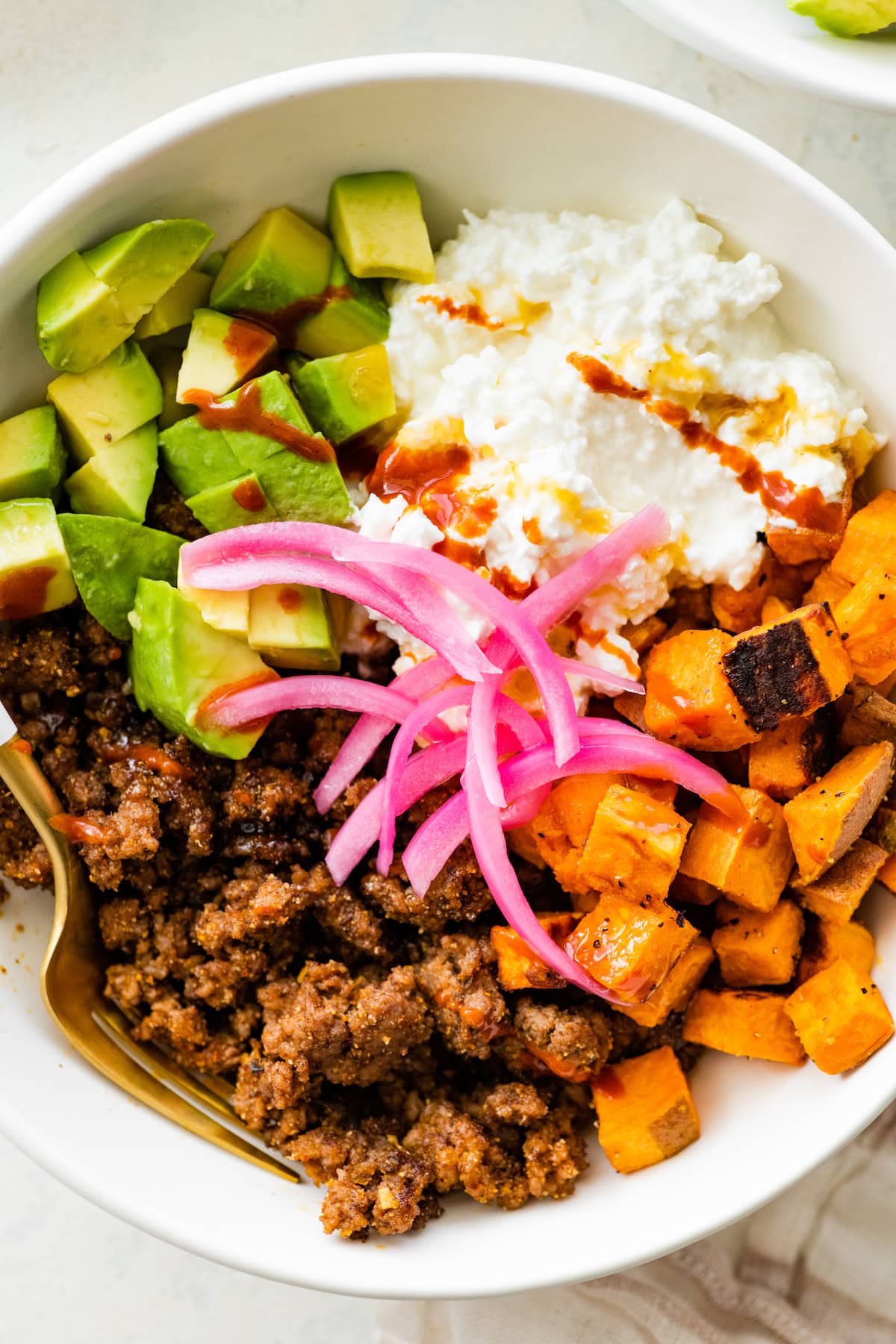 Ground beef, roasted sweet potatoes, cottage cheese and chopped avocado in a white bowl topped with honey and pickled onions and served with a fork.
