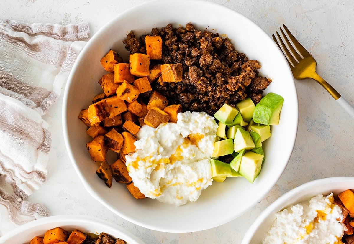 Ground beef, roasted sweet potatoes, cottage cheese and chopped avocado in a white bowl topped with a drizzle of honey.