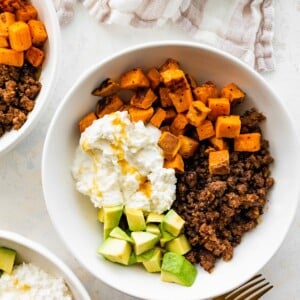 Ground beef, roasted sweet potatoes, cottage cheese and chopped avocado in a white bowl topped with a drizzle of honey.