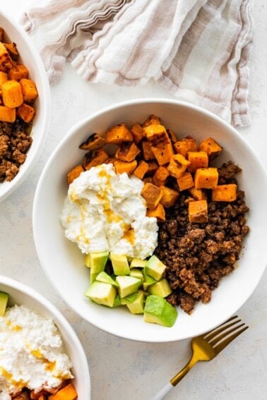 Ground beef, roasted sweet potatoes, cottage cheese and chopped avocado in a white bowl topped with a drizzle of honey.