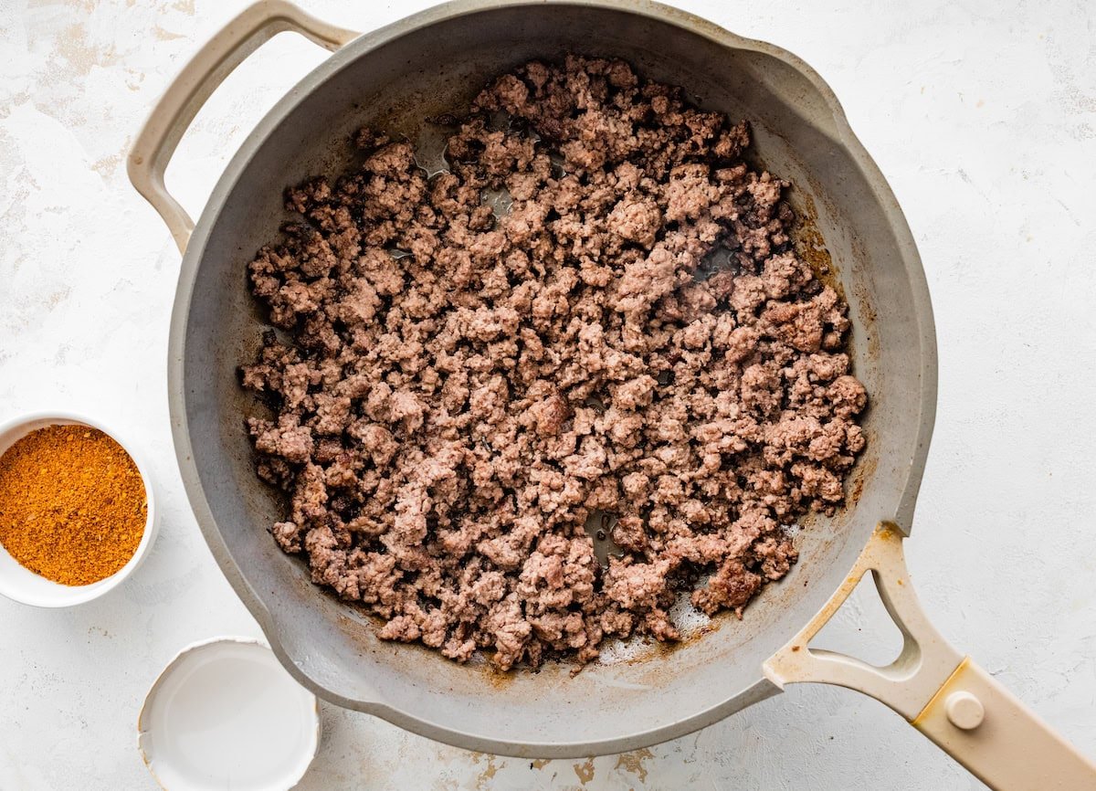 Cooked ground beef in a skillet with a small bowl of taco seasoning next to it.