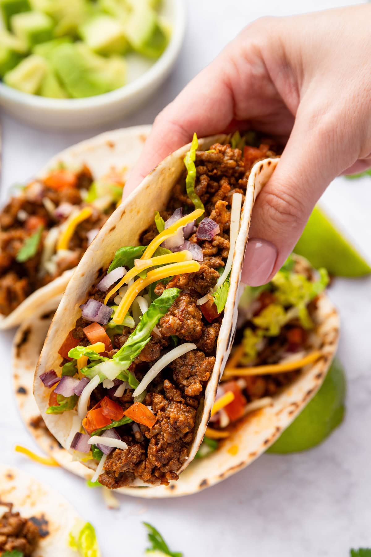 A woman's hand holding one ground beef taco that's topped with shredded cheese, onion, tomatoes and lettuce.
