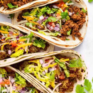 Five ground beef tacos in flour tortillas topped with lettuce, onion, tomato, cilantro and cheese.