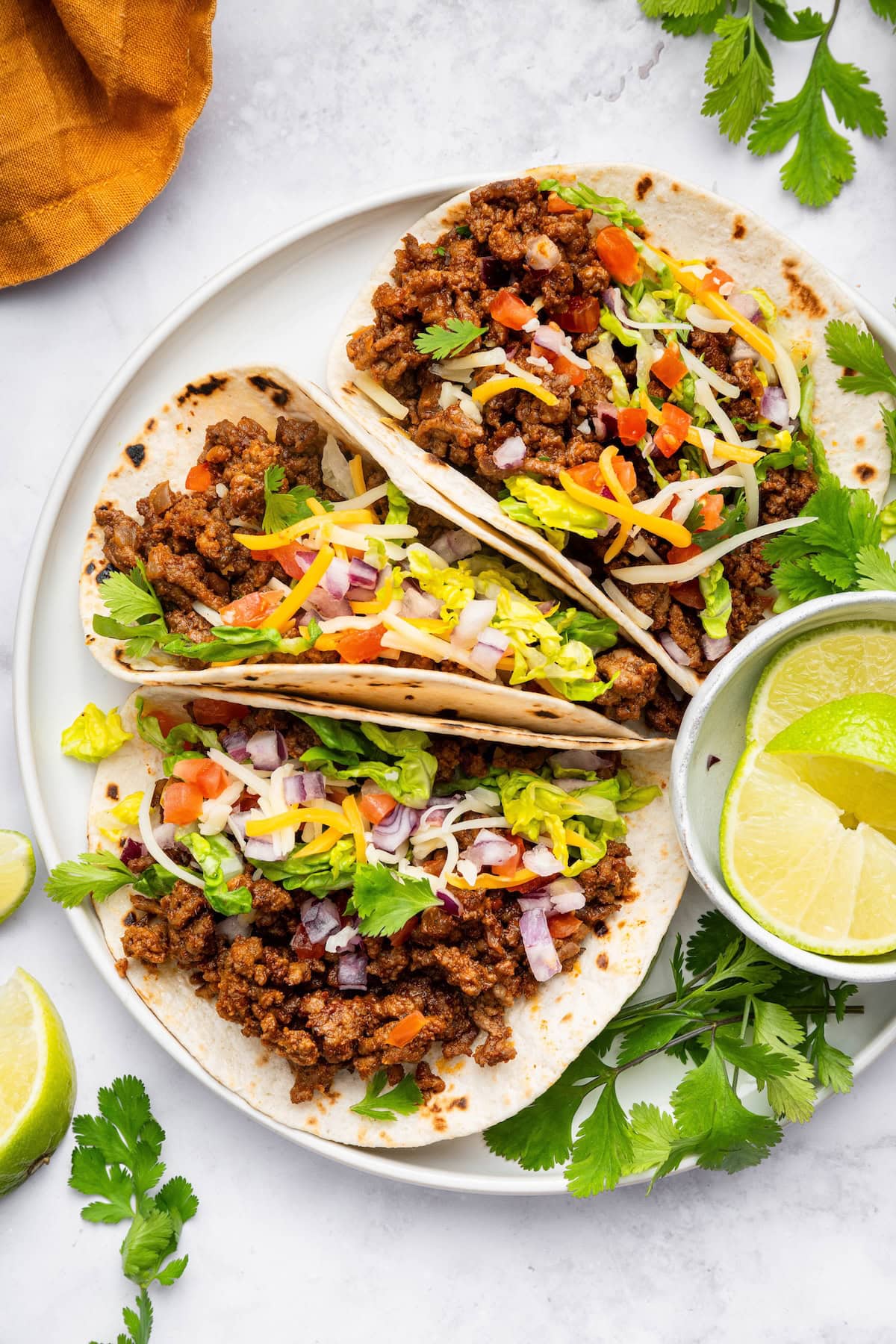 Three ground beef tacos in flour tortillas on a white plate topped with lettuce, onion, tomato, cilantro and cheese.