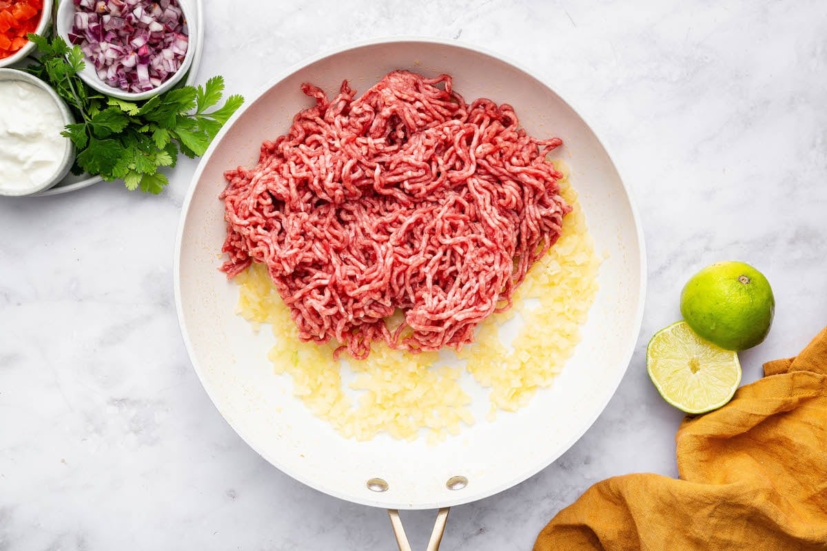 Raw ground beef added to a skillet with sautéed onion and garlic.