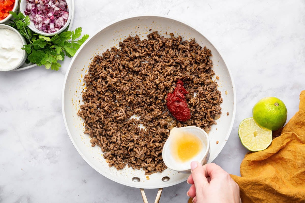 A woman's hand pouring broth into a skillet with a ground beef mixture and tomato paste.