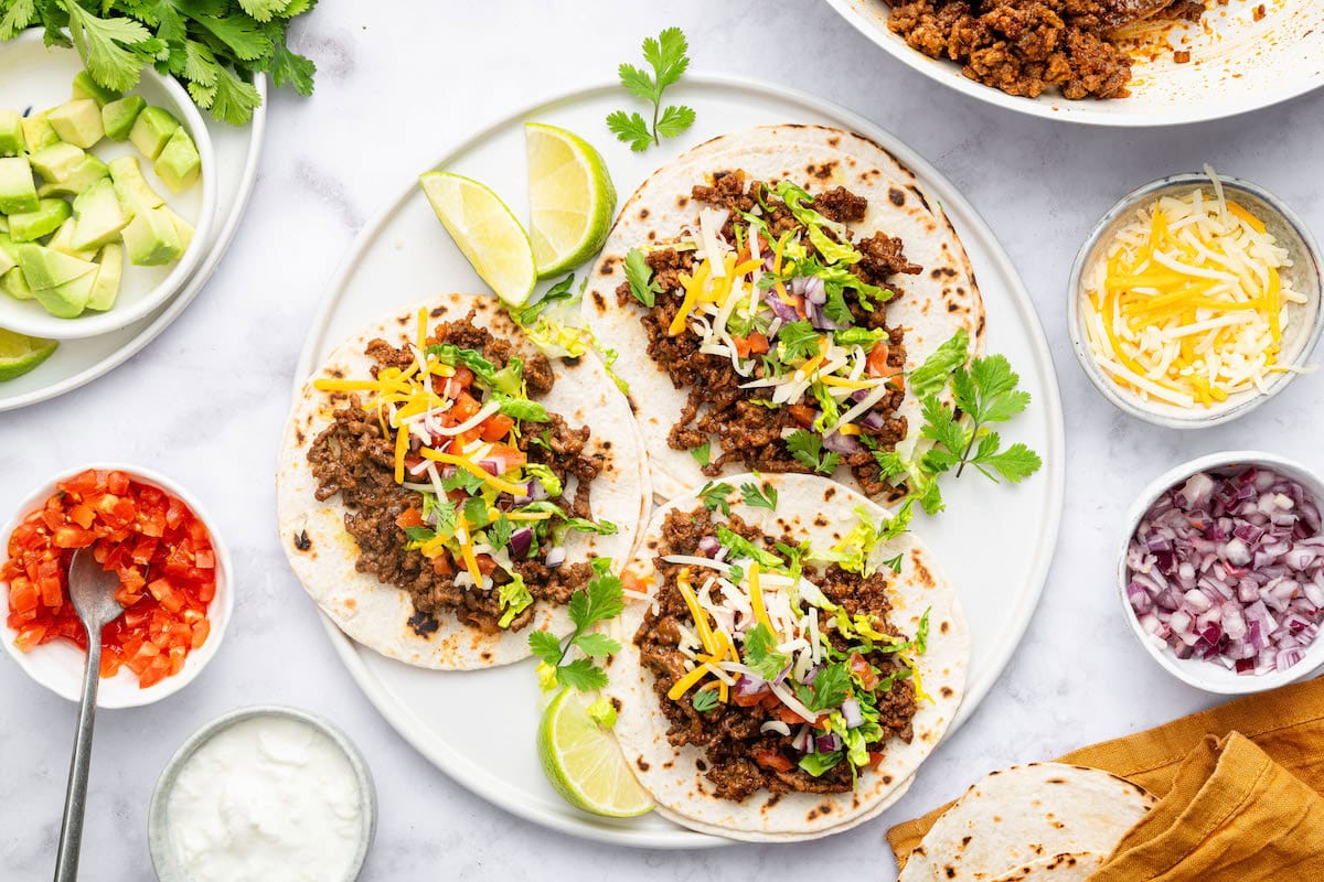 Three ground beef tacos in flour tortillas on a white plate topped with lettuce, onion, tomato, cilantro and cheese. Lime wedges are on the plate.