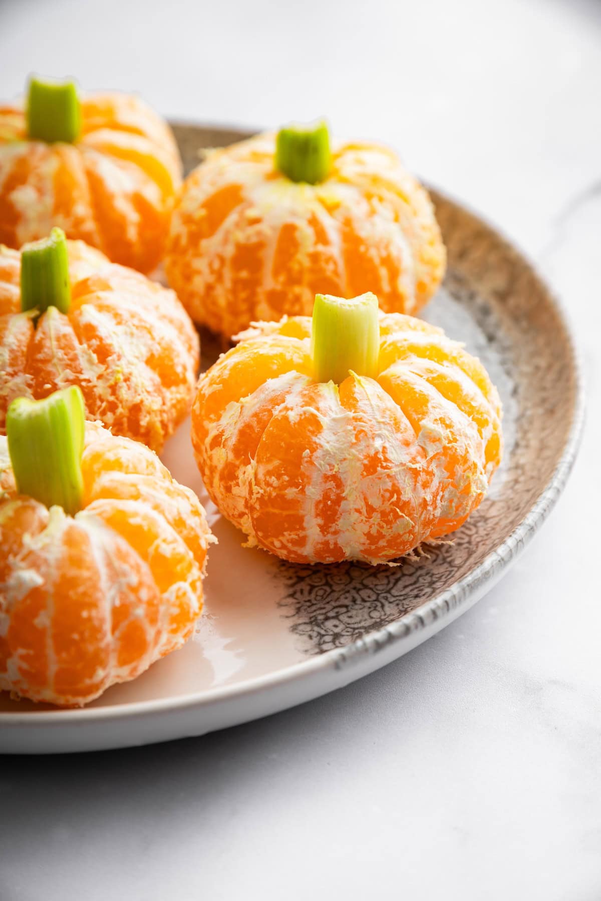 Peeled clementines topped with celery sticks to look like mini pumpkins on a small plate.