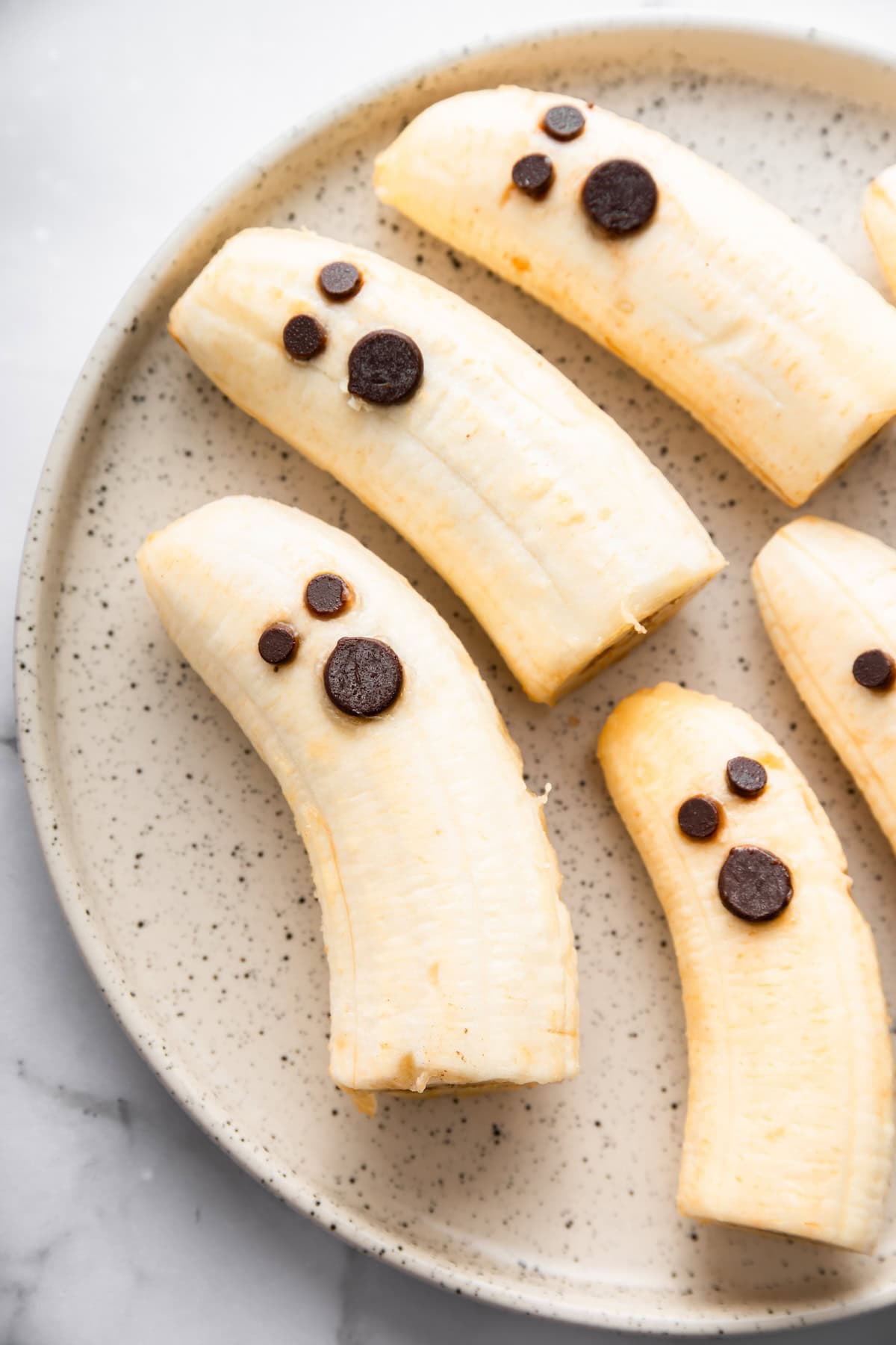 Banana halves decorated with mini and regular chocolate chips to look like spooky ghosts on a white plate.