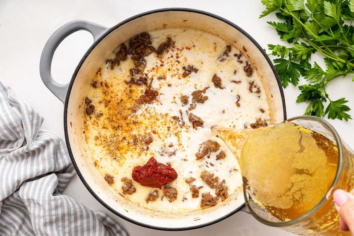 Broth is poured into a large pot of ground beef, tomato paste, seasonings, onion, and garlic.