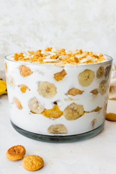 A glass trifle dish filled with healthy banana pudding. Layers of fresh banana slices and wafer cookies are visible in between the yogurt and coco whip.