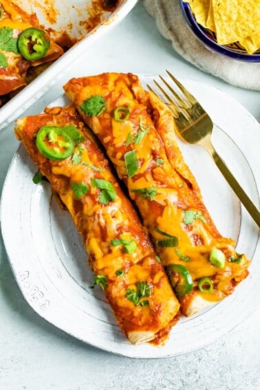 Two chicken enchiladas on a plate topped with cheese and cilantro with a fork on the plate.