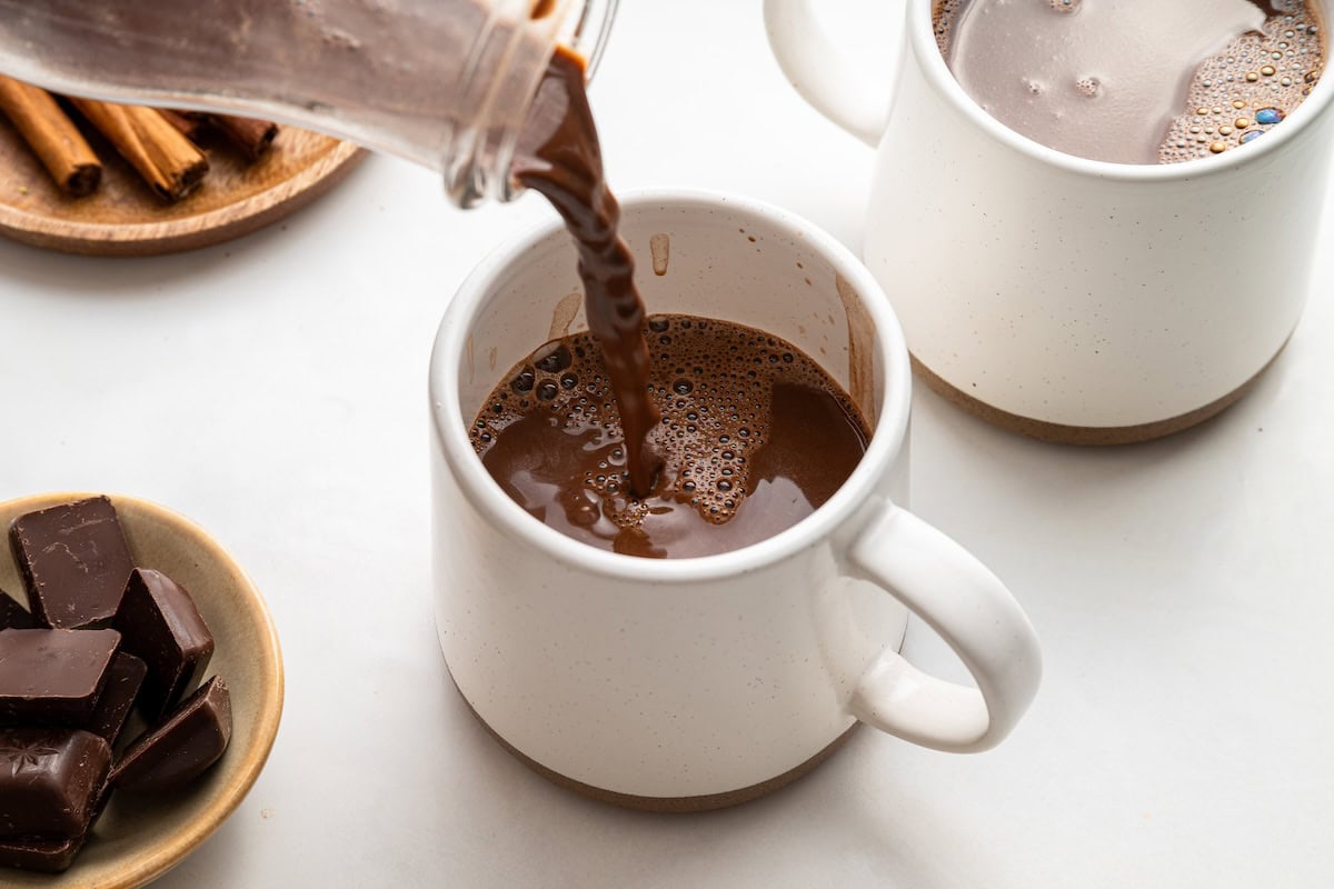 Hot chocolate being poured into a white mug with another mug behind it.