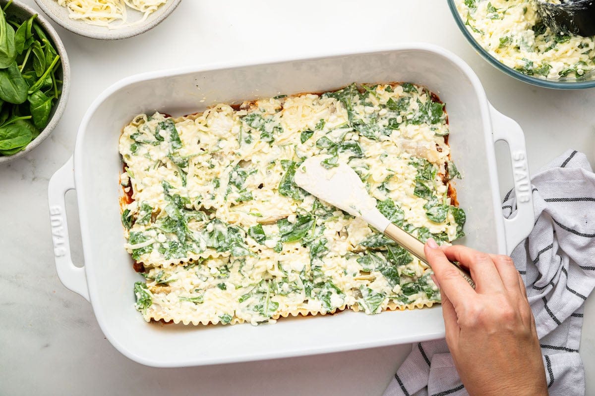 A woman's hand uses a spatula to spread a cheese layer on lasagna noodles.