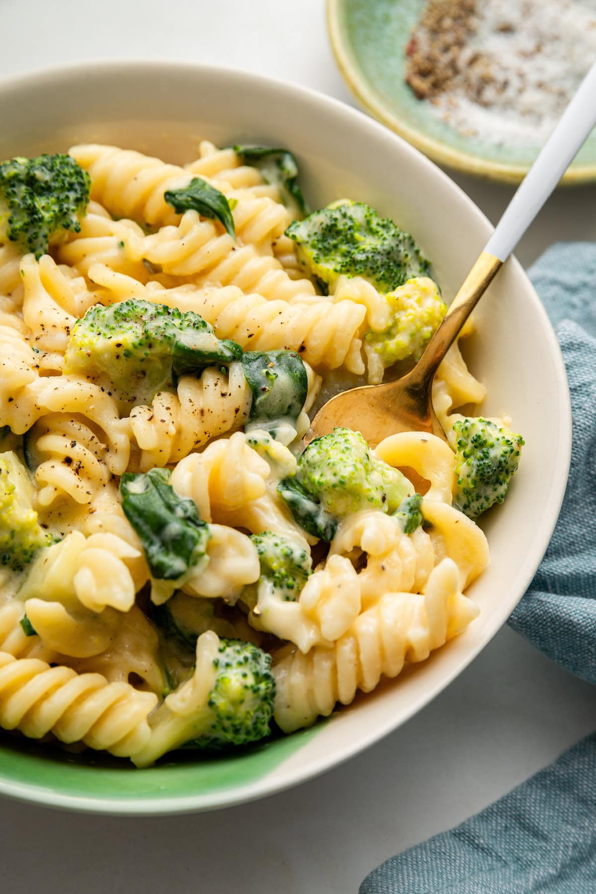 One bowl of healthy mac and cheese served with a gold fork. The bowl is filled with rotini pasta, broccoli and spinach all coated in a cheese sauce and topped with fresh pepper.