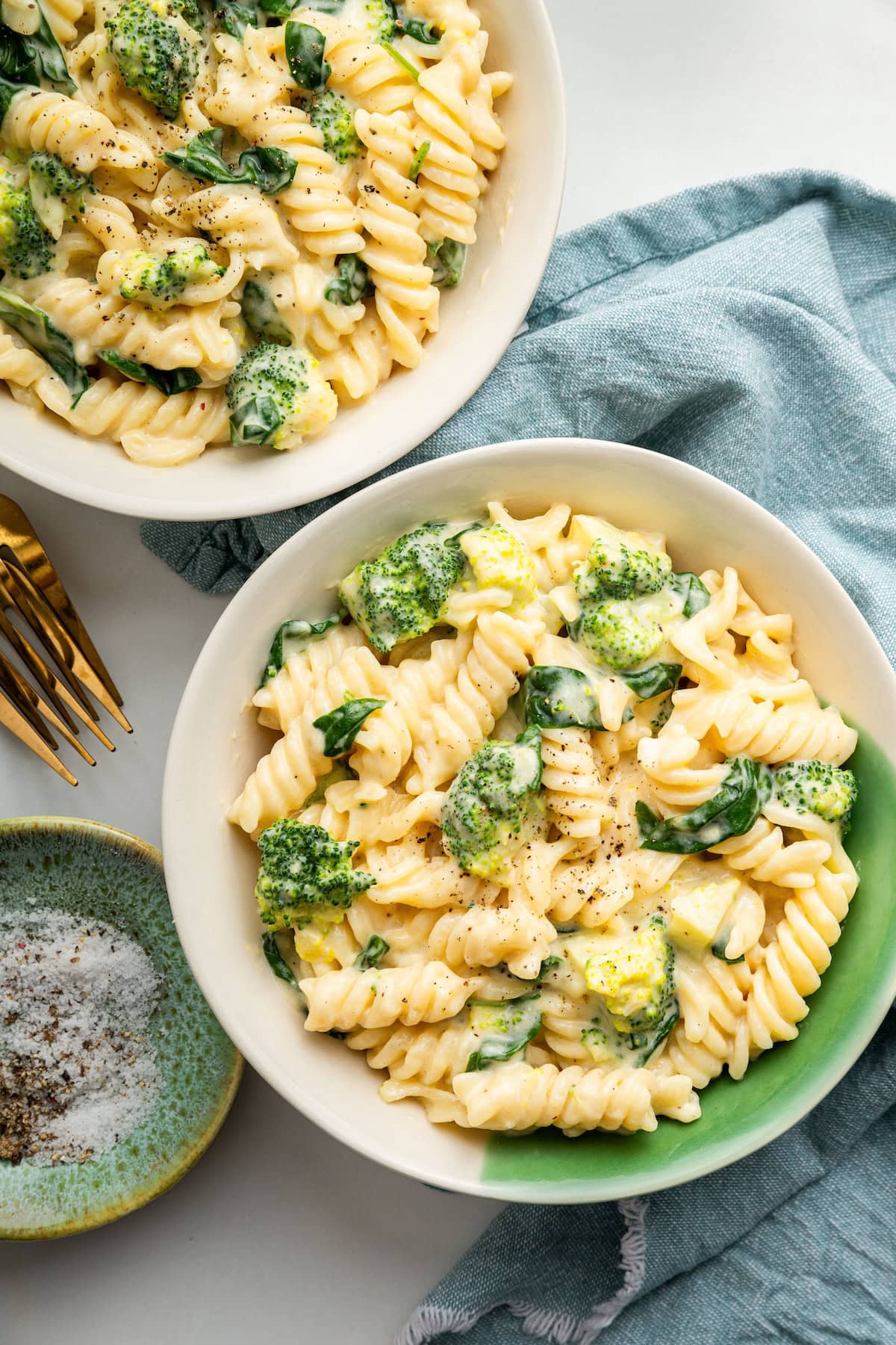 Two bowls of healthy mac and cheese. The bowls are filled with rotini pasta, broccoli and spinach all coated in a cheese sauce.