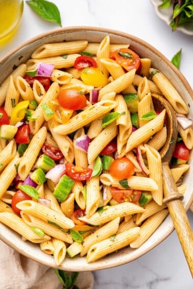 A pasta salad in serving bowl with cherry tomatoes, cucumber, red onion and fresh basil. There is a wooden serving spoon in the bowl.
