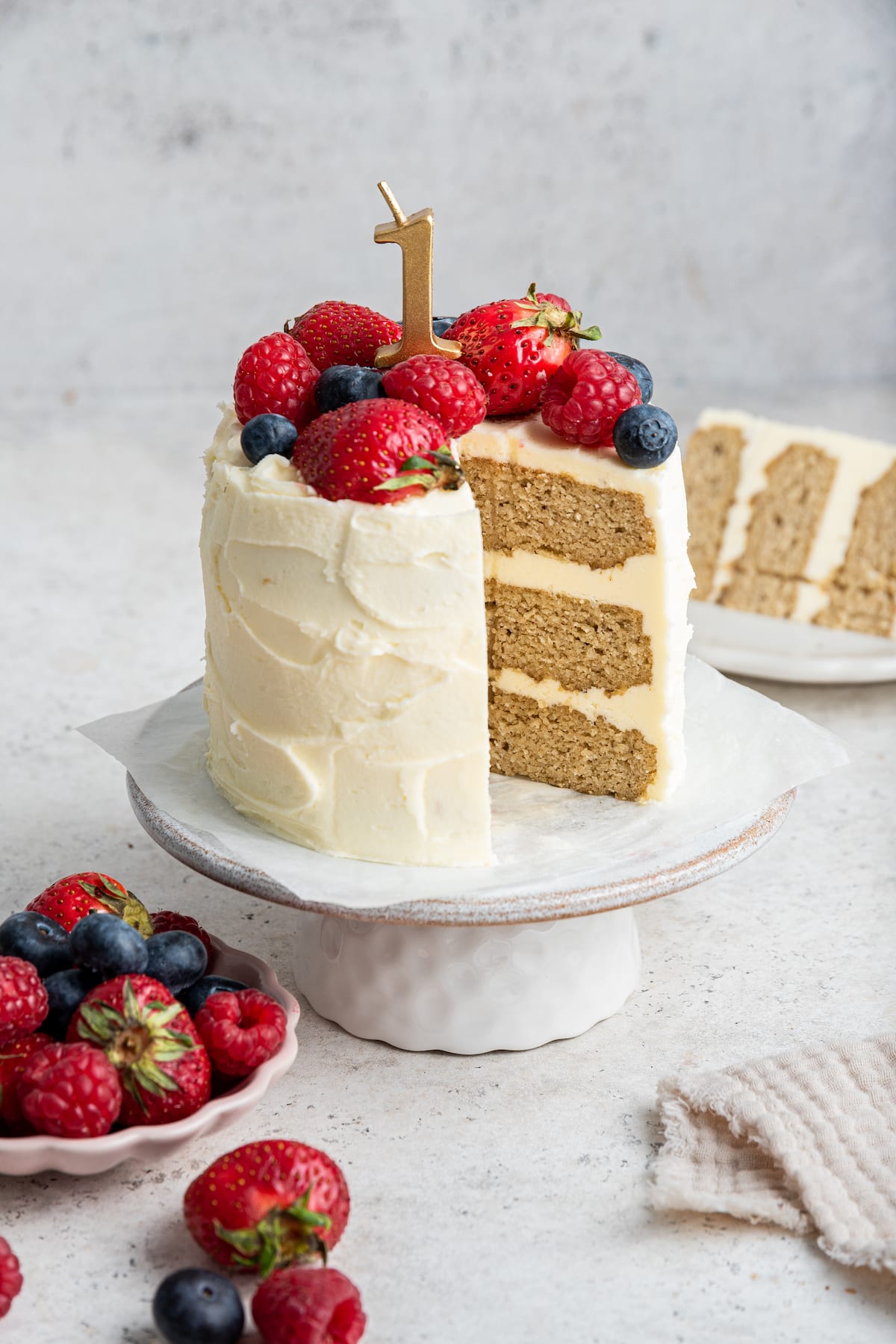 A frosted smash cake with a one candle topped with fresh berries sitting on a cake stand. One slice of the cake has been cut out of it.