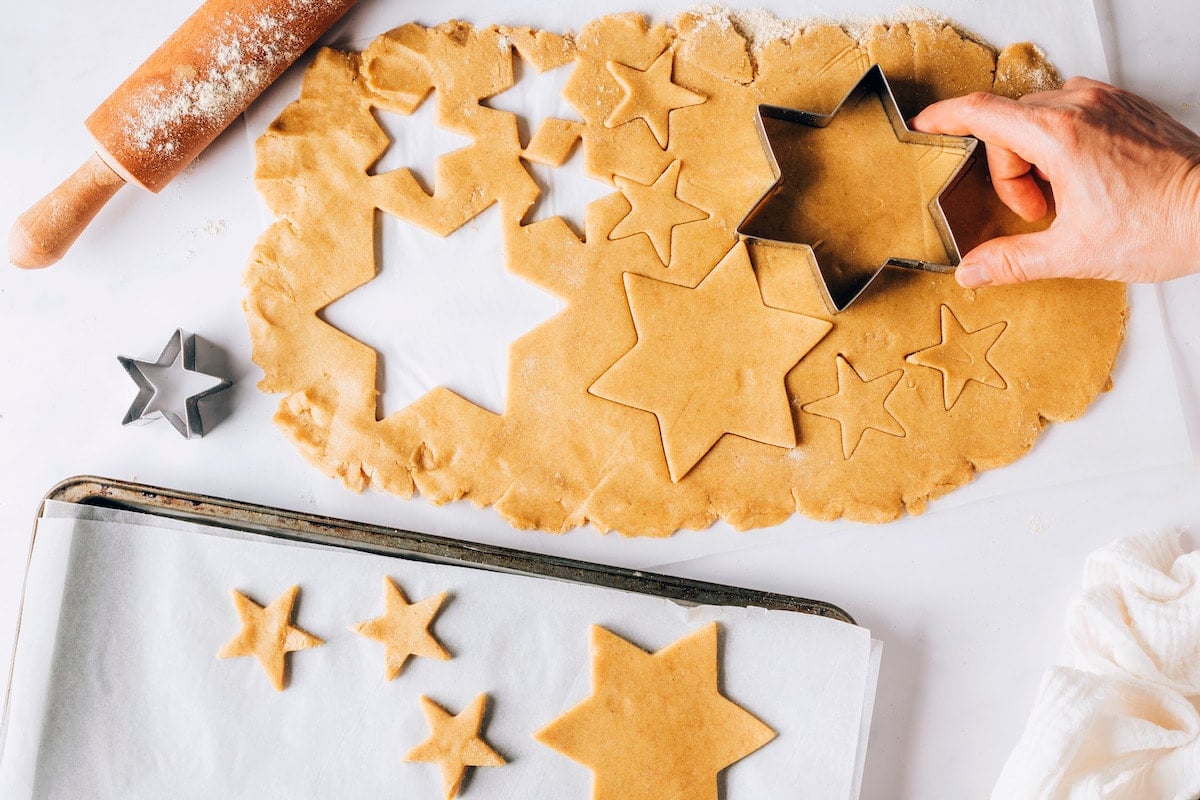 A hand using a star shaped cookie cutter to cut out cookies from rolled dough. The cookies are being placed onto a parchment lined baking sheet.