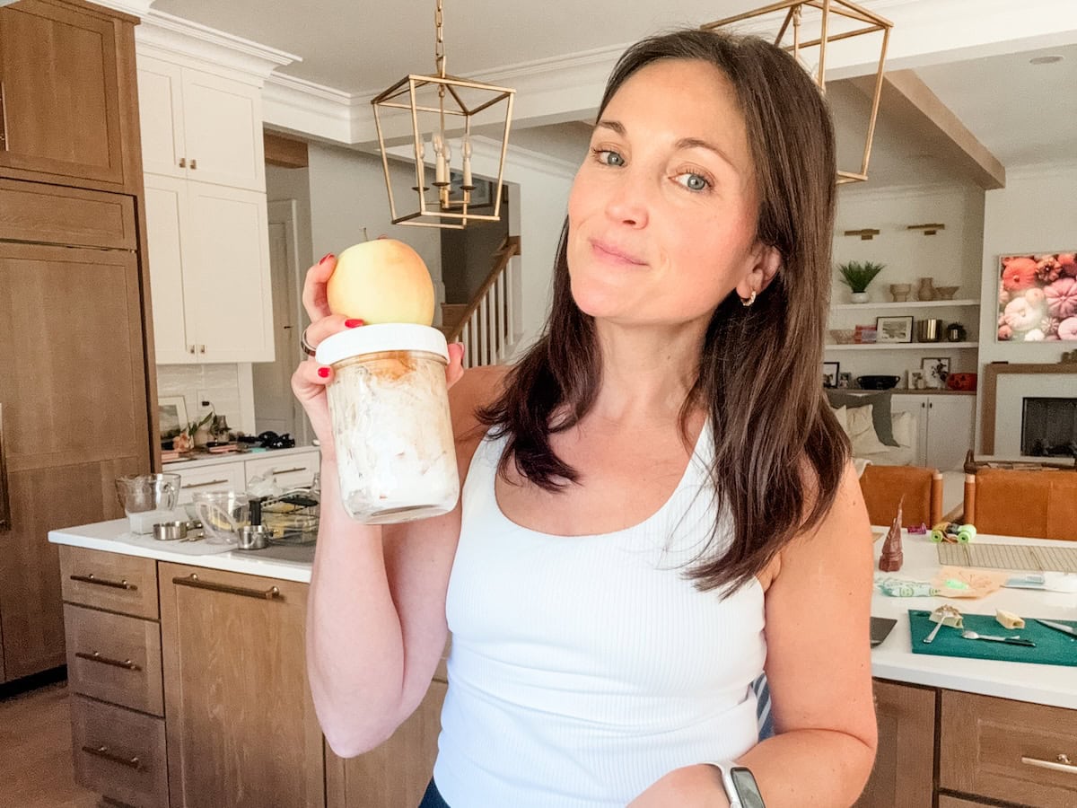 A woman holding a sealed glass mason jar that contains high protein apple salad, with a full apple on top of the jar.