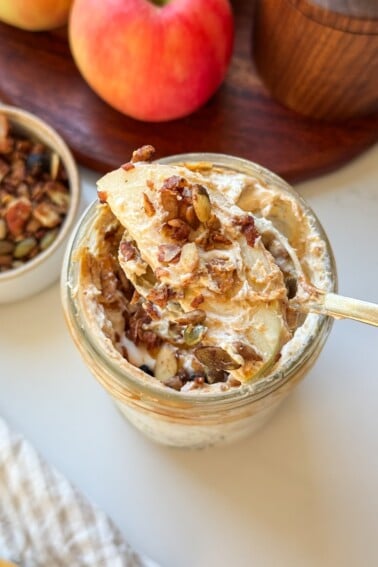 A spoonful of high protein apple salad with an apple slice and granola, held over a mason jar.