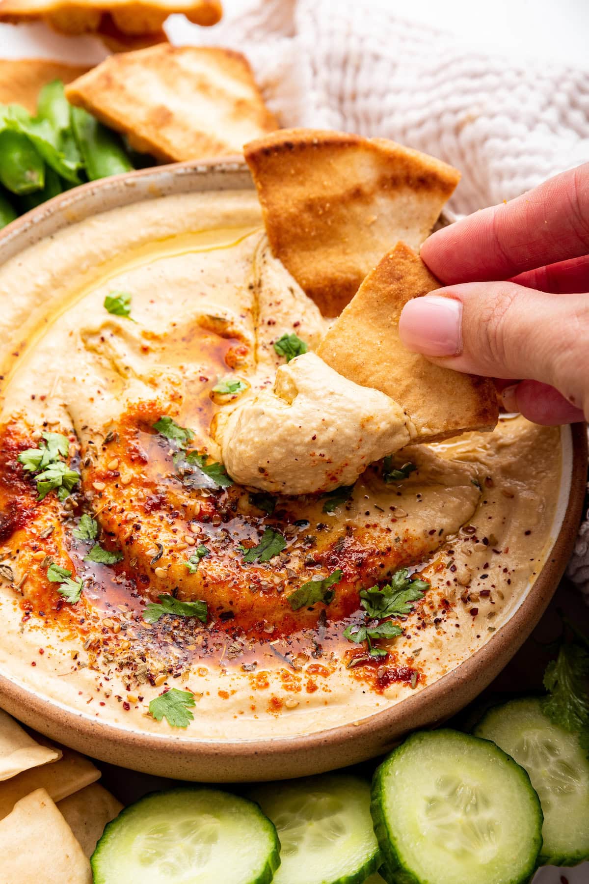 A woman's hand dips a pita chip in a bowl of creamy hummus topped with herbs and spices.