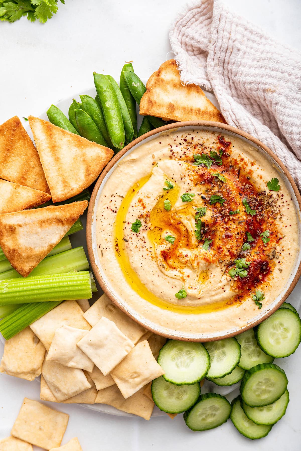 A bowl of creamy hummus topped with herbs and spices. Near the bowl are pita chips and fresh vegetables.