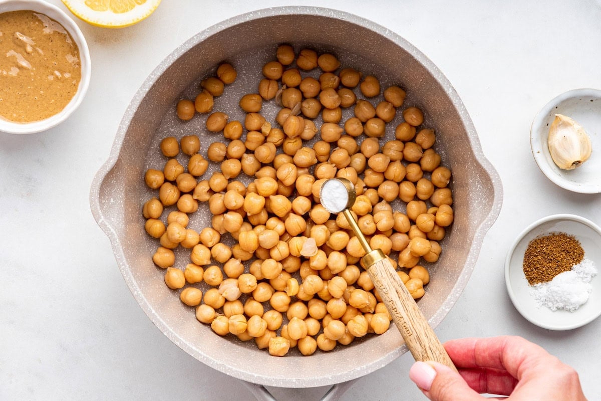 A woman's hand adds a teaspoon of baking soda to a large pot of garbanzo beans.