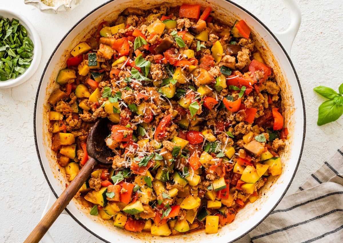 Large skillet filled with cooked ground turkey, vegetables and tomato sauce topped with basil and fresh parmesan cheese. There is a wooden spoon in the mixture.