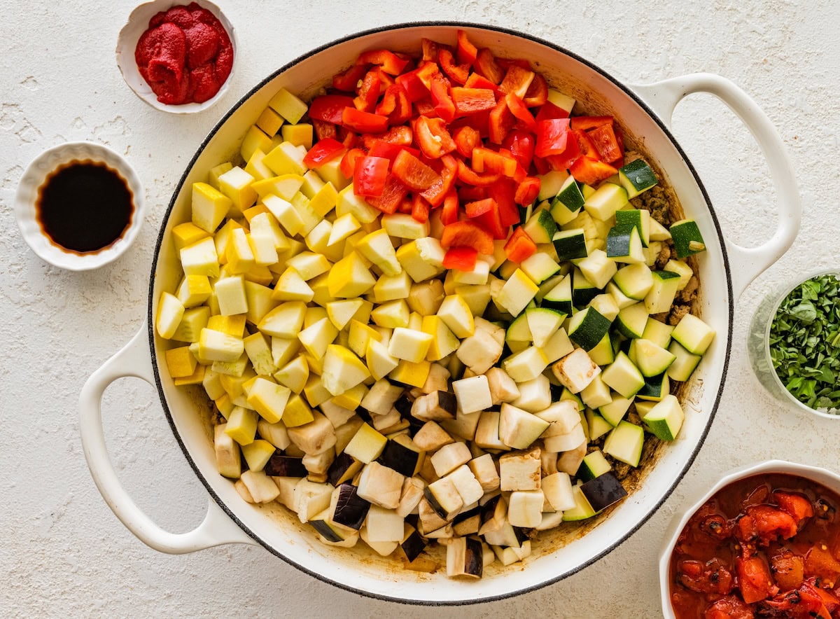 Chopped zucchini, yellow squash, red bell pepper, and eggplant added to a white skillet.