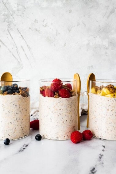 Three glass cups of kefir overnight oats. One cup is topped with fresh blueberries, another with fresh raspberries, and the last with fresh pineapple.