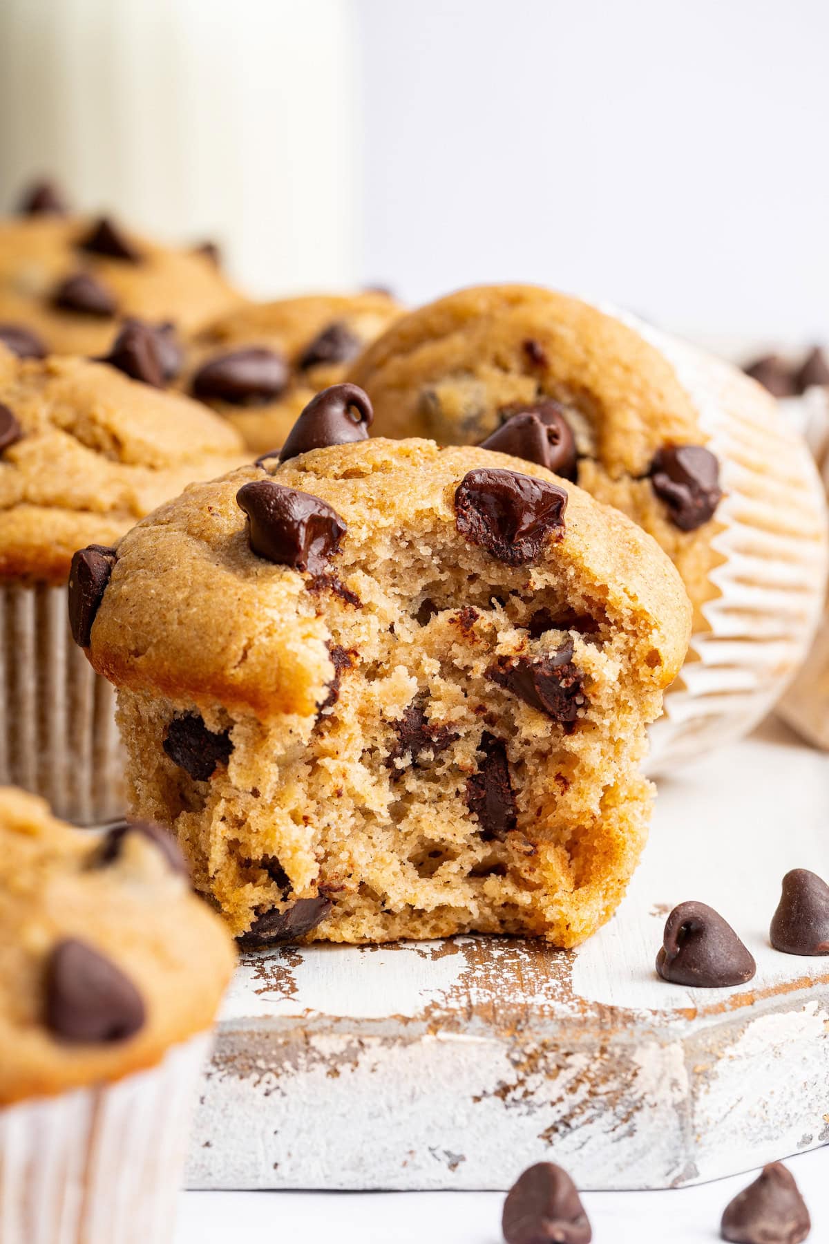 A Kodiak cake muffin with a bite taken out, showing the soft inside with melted chocolate chips. Other muffins and chocolate chips are in the background.