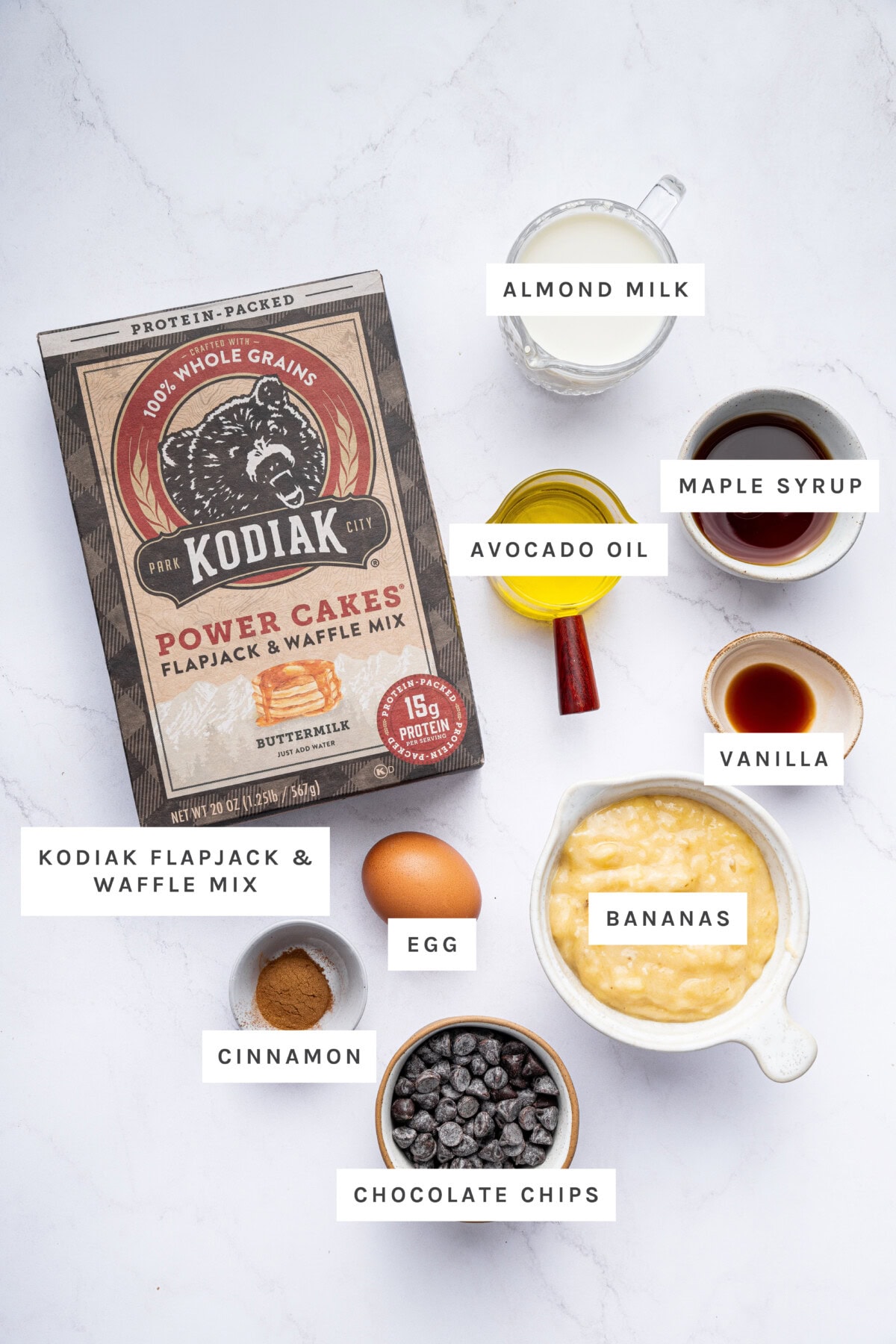 Ingredients measured out to make Kodiak Cake Muffins: flapjack & waffle mix, almond milk, maple syrup, avocado oil, vanilla, bananas, egg, cinnamon, and chocolate chips.