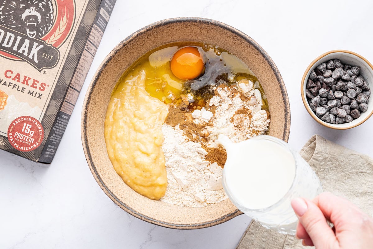 A bowl with ingredients for Kodiak cake muffins, including mashed banana, an egg, Kodiak pancake mix, spices, and milk being poured in.
