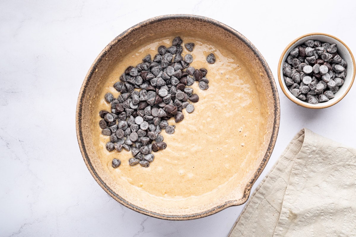 A bowl of mixed muffin batter with chocolate chips on top, ready to be folded in. A small bowl of extra chocolate chips is beside it.