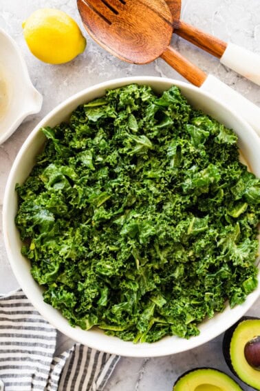 Massaged kale salad in a large white serving bowl with a lemon and wooden salad servers beside it.