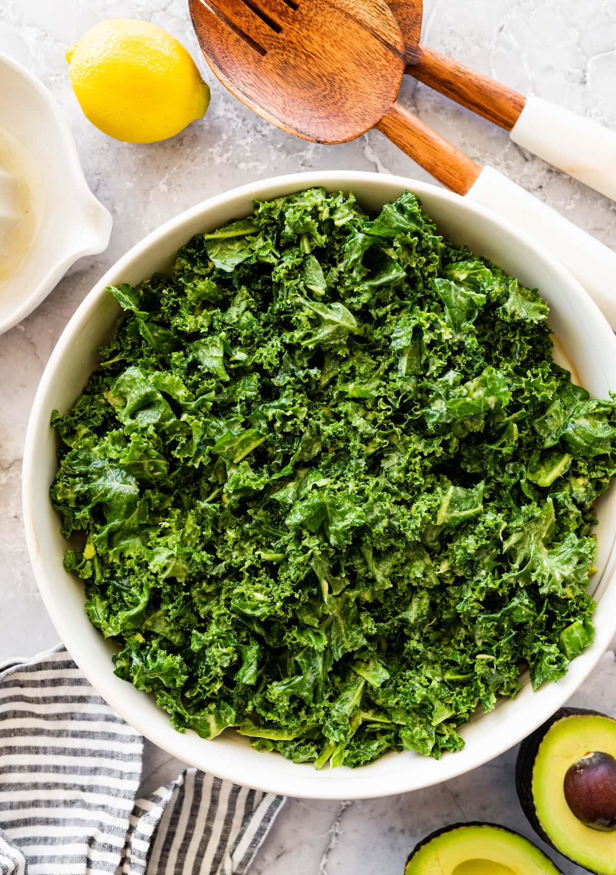 Massaged kale salad in a large white serving bowl with a lemon and wooden salad servers beside it.