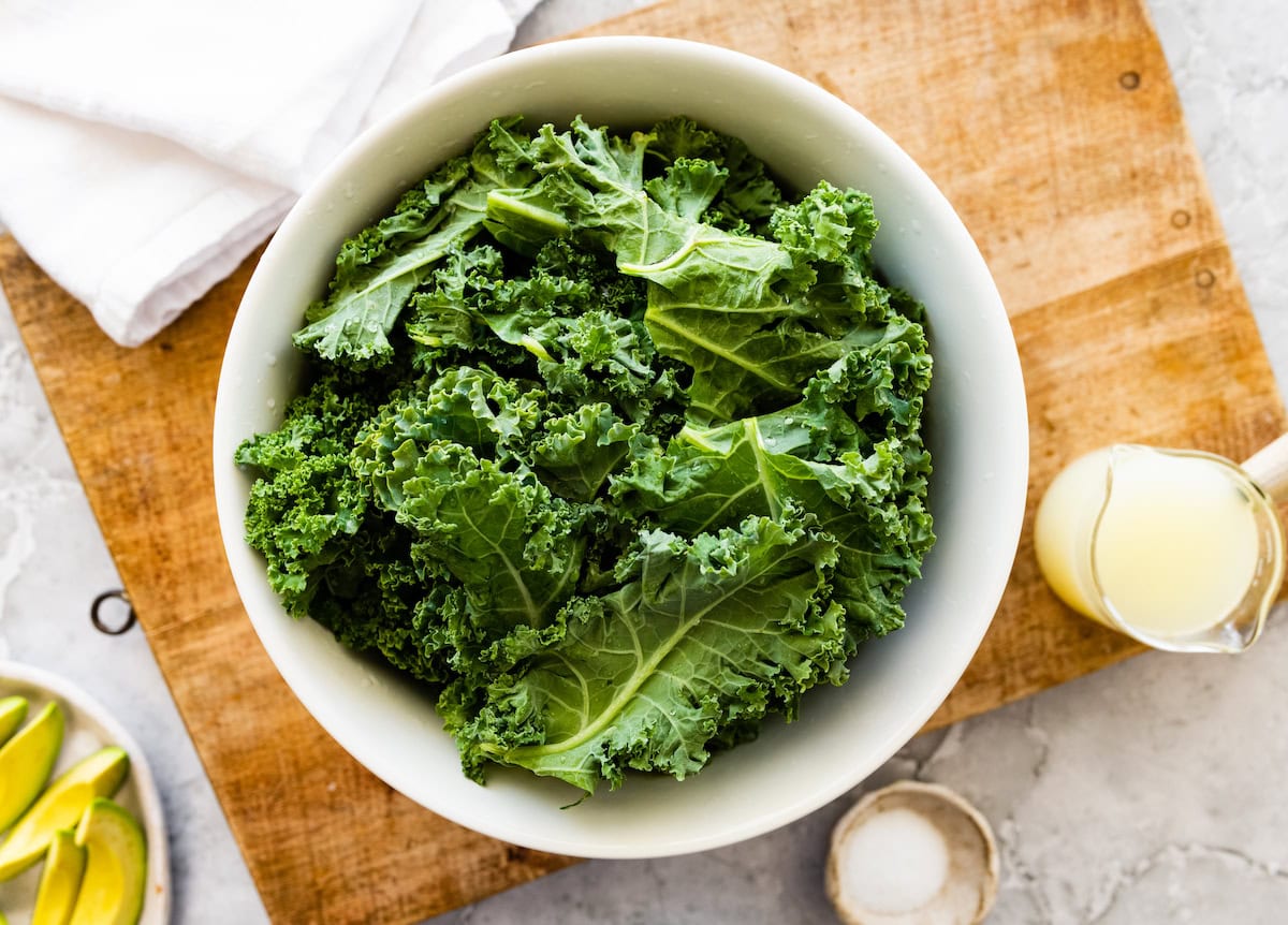 Fresh curly kale leaves in a white bowl on a wooden board with lemon juice and sliced avocado nearby.