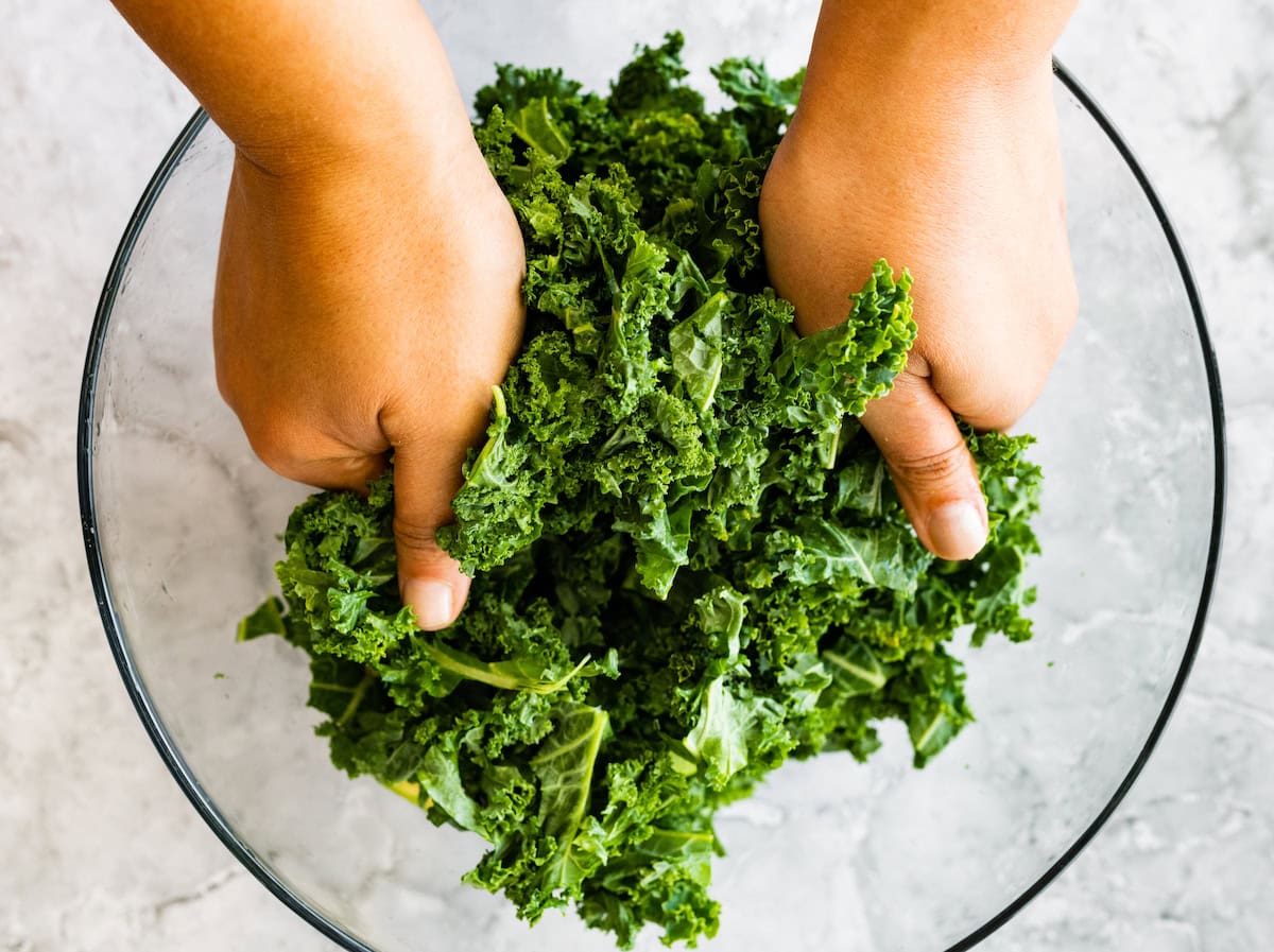 Two hands massaging fresh chopped kale in a glass mixing bowl.