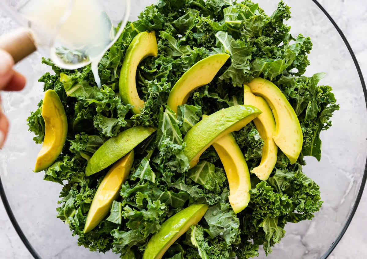 Sliced avocado on top of fresh kale in a mixing bowl with lemon juice being poured in.