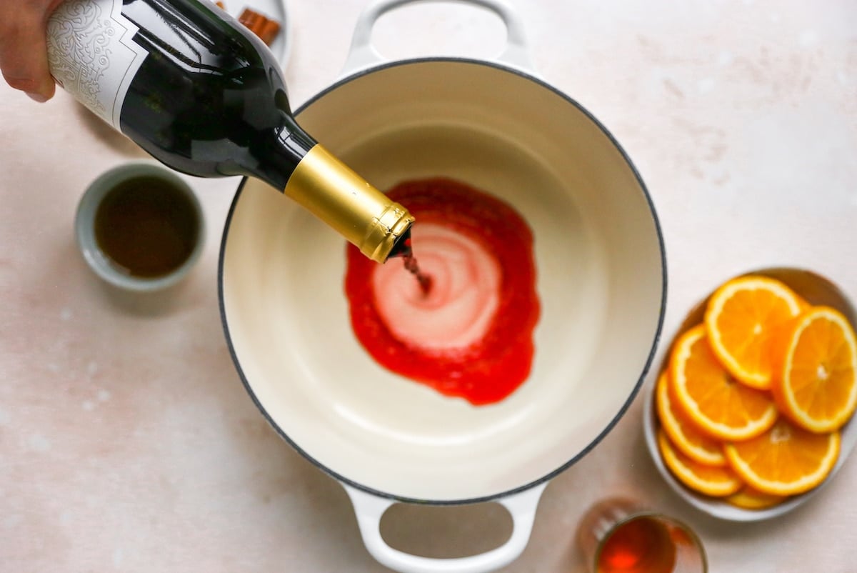 A large pot being filled with red wine. Additional ingredients for mulled wine are displayed around the pot.