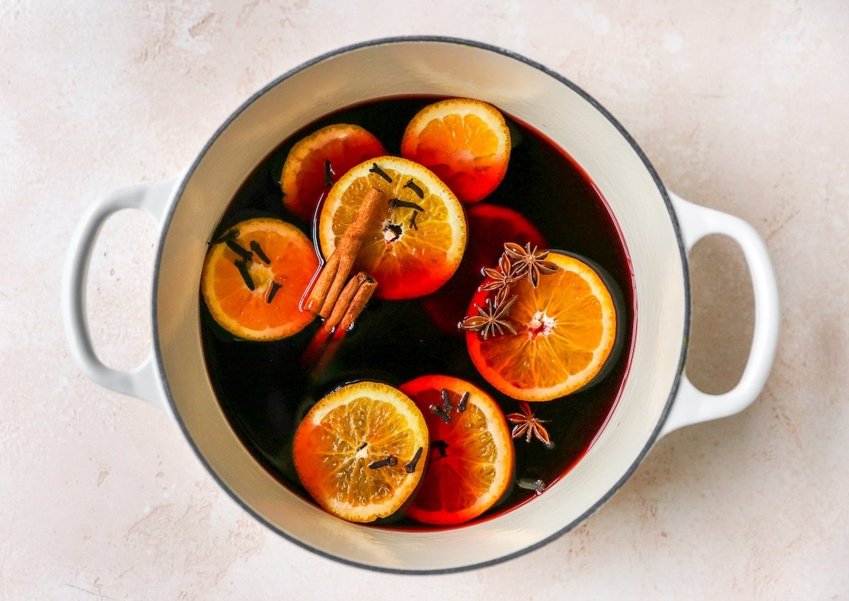 A large pot containing ingredients for mulled wine: red wine, brandy, maple syrup, orange slices, cinnamon sticks, cloves and star anise.