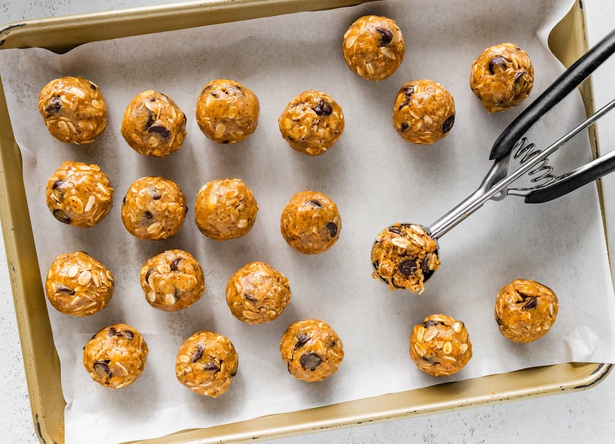20 energy balls on a parchment-lined baking sheet. A cookie scoop is filled with the dough to make one of the energy balls.
