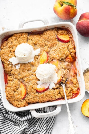 Overhead view of a baked peach cobbler with golden brown topping and two scoops of vanilla ice cream in a white square dish.