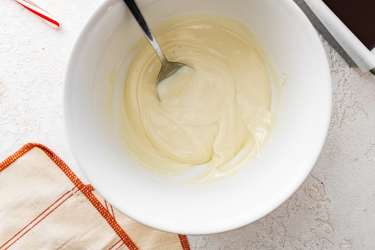 Melted white chocolate in a white mixing bowl with a silver spoon.