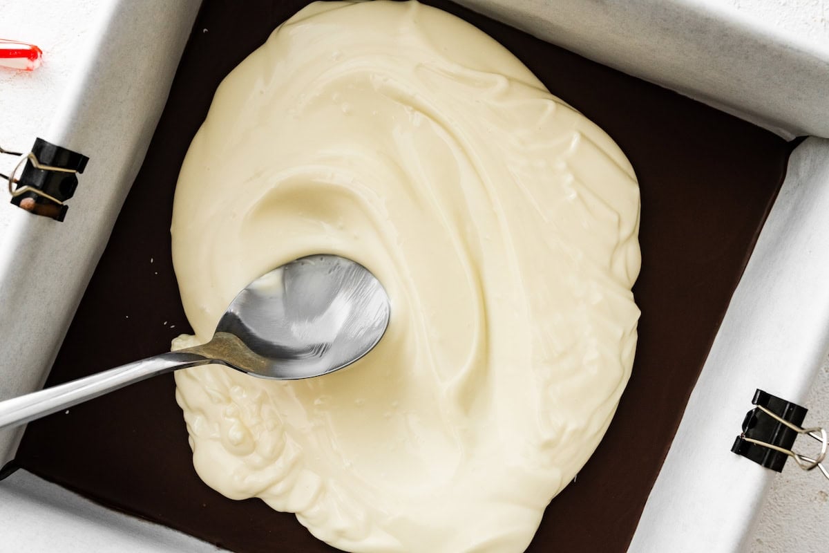 The melted white chocolate being spread on top of the chilled dark chocolate layer with a silver spoon.