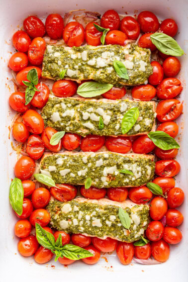Four salmon filets covered in pesto and surrounded by cherry tomatoes in a baking dish.
