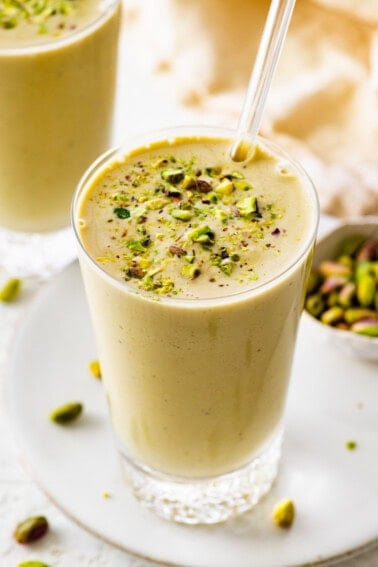 One glass filled with a pistachio smoothie topped with crushed pistachios. There is a clear straw in the glass and another smoothie in the background.