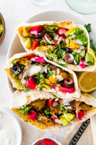 Three halves of chicken shawarma pita pockets stacked in a bowl, filled with veggies, olives, pickled turnips, and topped with yogurt sauce and fresh parsley.